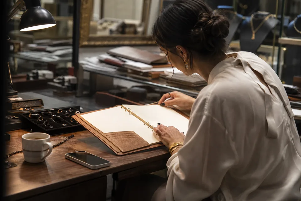 Jeweler reviewing her client book and phone at a desk in the back office before calling clients